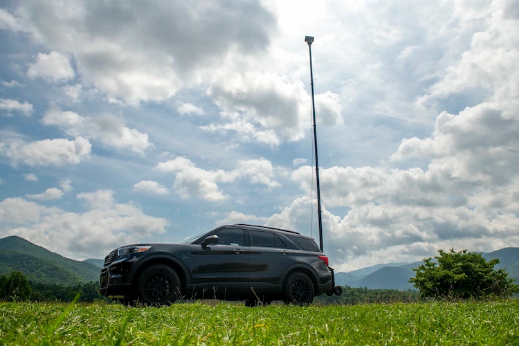 Modern black SUV with a high-reaching surveillance camera setup, stationed in a vibrant green field with mountains and a cloudy sky in the backdrop.