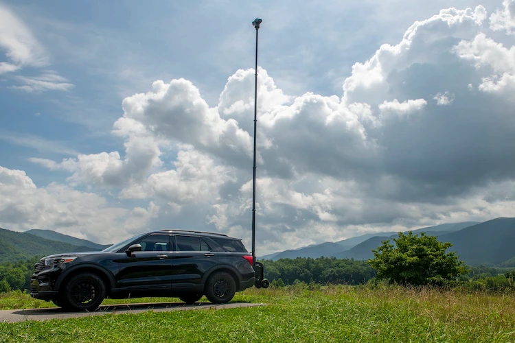 Black SUV with a telescopic surveillance camera mast, parked in a grassy field with mountain ranges and puffy clouds in the background.