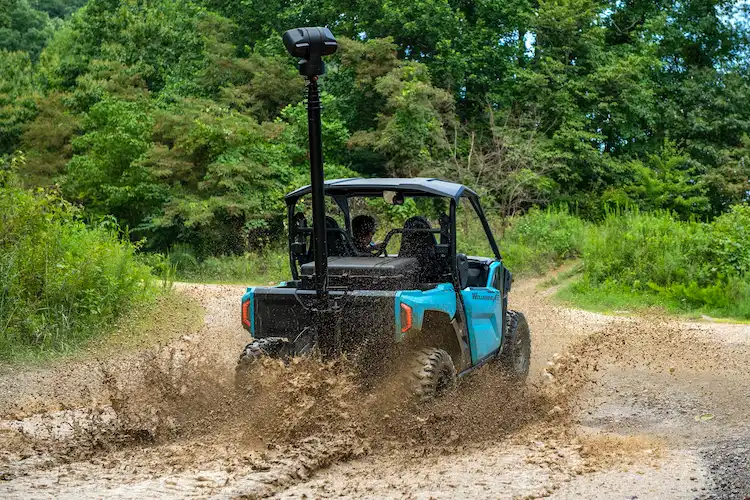 Off-road utility vehicle with a mounted surveillance mast splashing through a muddy trail surrounded by lush greenery.