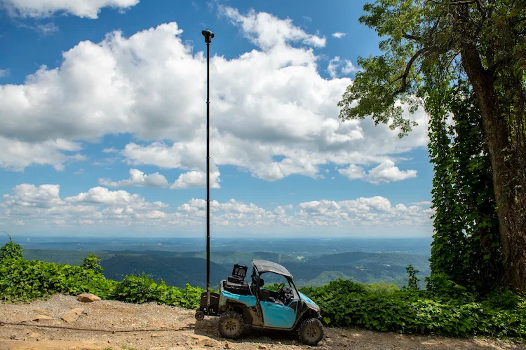Blue off-road vehicle equipped with a high surveillance camera, parked on a mountain edge with panoramic views of lush landscapes and cloudy skies.