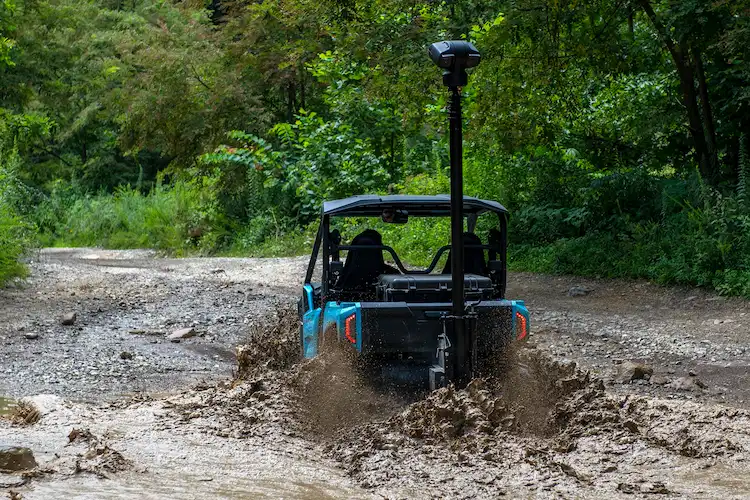 Off-road utility vehicle with a mounted surveillance mast splashing through a muddy trail surrounded by lush greenery.