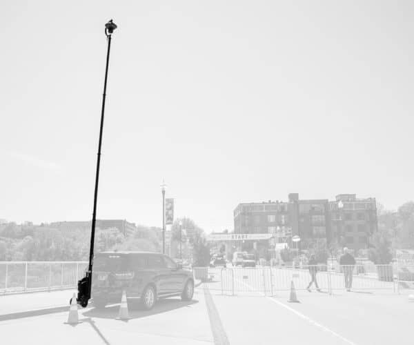 A mobile surveillance unit with a raised camera tower on an SUV, positioned at a public event with barricades and people walking in the background.