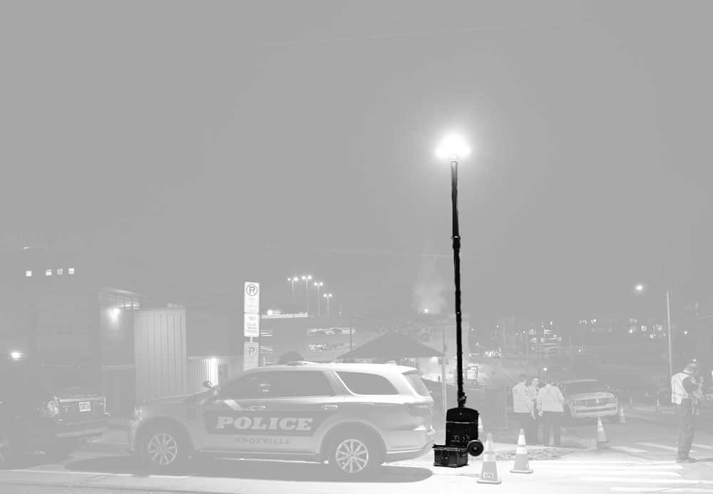 Grayscale image of a portable light tower on a small trailer, with extended mast and LED lights, parked outdoors near trees and power lines.