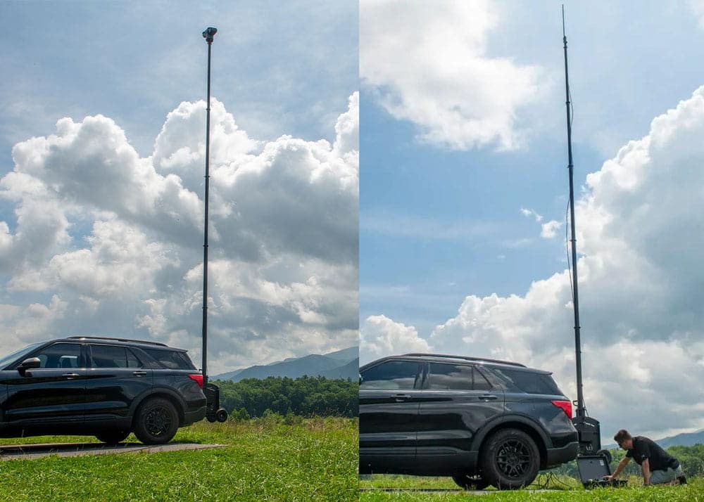 Telescopic mast deployed on a mobile surveillance tower beside an antenna mast deployed on a vehicle in an open outdoor setting.
