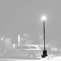 Grayscale image of a portable light tower on a small trailer, with extended mast and LED lights, parked outdoors near trees and power lines.